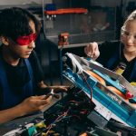 Two students work together on a robotics project in a workshop, wearing safety gear.